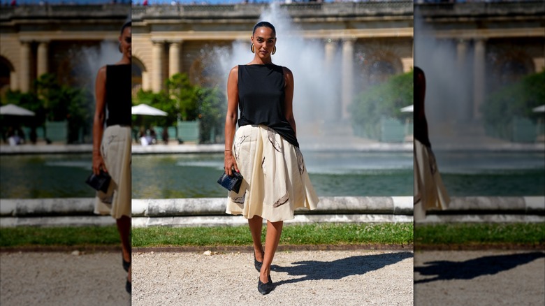 A woman in a black boatneck sleeveless top and a pleated white skirt standing in front of a fountain.