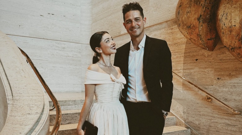 Sarah Hyland in a creme colored gown and a slicked back ponytail standing next to Wells Adams in a black suit
