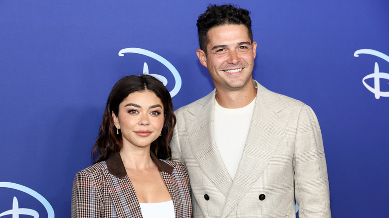 Sarah Hyland in a brown plaid blazer posing next to Wells Adams in a cream blazer