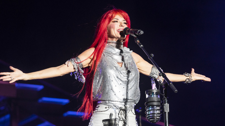 Shania Twain performs on stage while wearing a bright red wig and a metallic silver halter top.