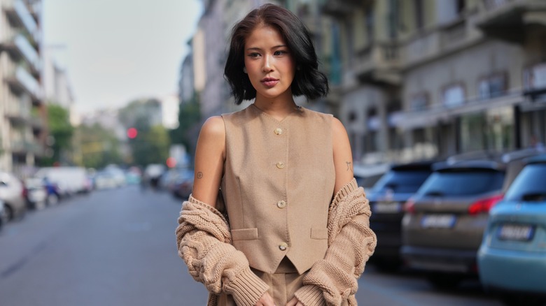A woman wearing a tan button-up longline vest and a matching sweater off the shoulders.