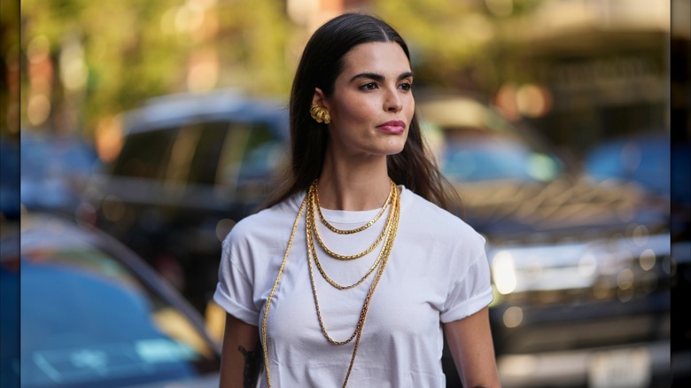 A woman wearing a white t-shirt with rolled sleeves and several gold chain necklaces.
