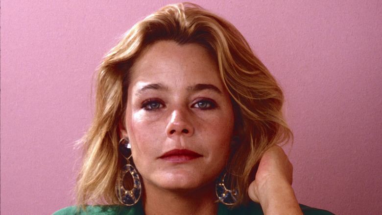 Susan Dey wearing a green top and statement earrings posing in front of a pink backdrop.