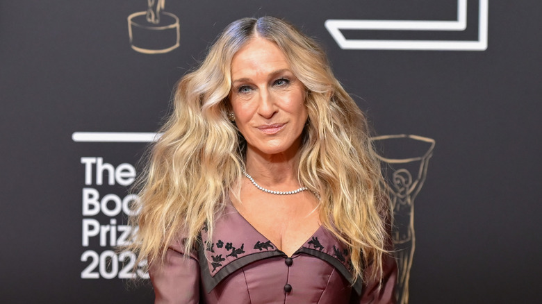 Sarah Jessica Parker posing at an event wearing a maroon gown with wavy hair and a delicate necklace.