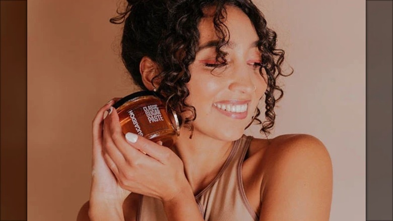 Sugardoh founder Aliyah Marandiz posing with a jar of hair removal paste by her face.