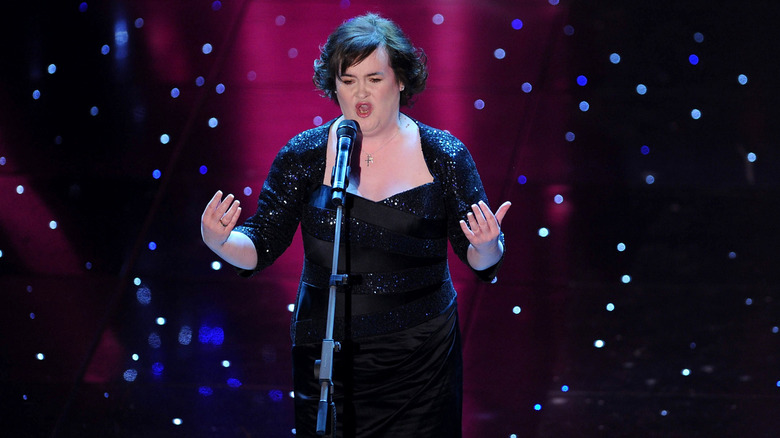 Susan Boyle sings into a microphone wearing a black sequin dress