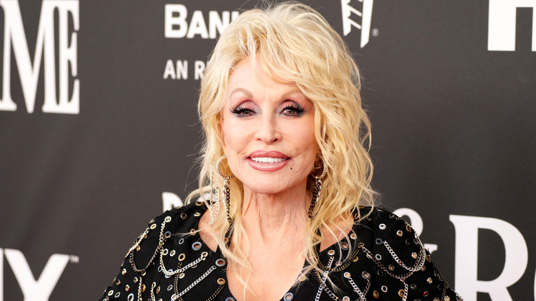 Dolly Parton smiles on the red carpet in full flam while standing in front of a black and white backdrop.