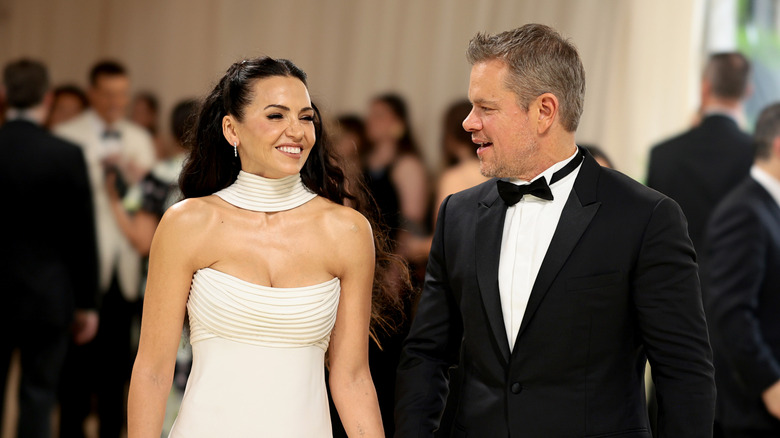 Luciana Barroso in a white gown and Matt Damon in a tuxedo smile at each other on the red carpet.
