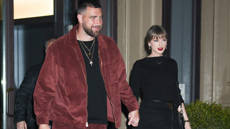 Travis Kelce wear a red jacket pictured with Taylor Swift wearing an all-black outfit