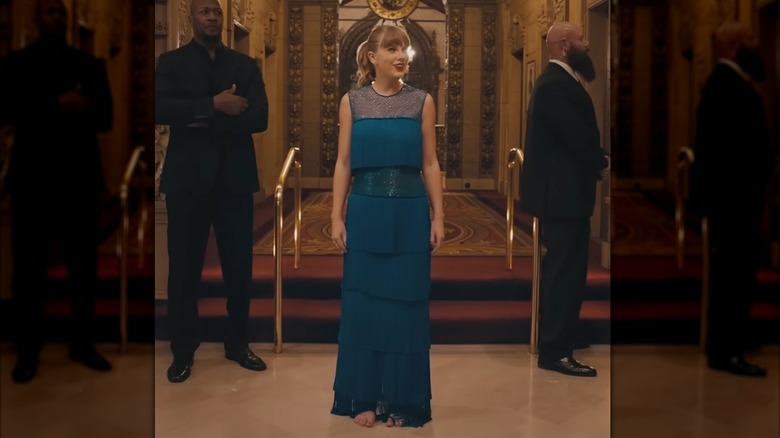 Swift in the hallway of a grand hotel. She is wearing a blue dress with several layers of fringe. Her hair is back in a ponytail.