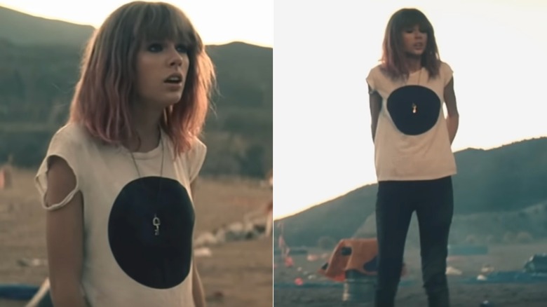 Taylor Swift stands alone in her "I Knew You Were Trouble" music video. She is wearing a blonde wig with pink ends, black skinny jeans, and a ripped white t-shirt with a black circle design.