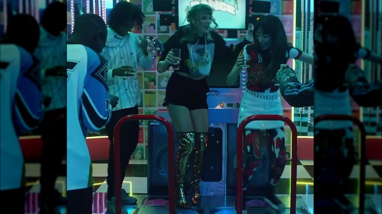 Taylor Swift plays a dance game at an arcade. She is wearing a cropped graphic hoodie, black track shorts, and a pair of over-the-knee gold sequin boots.