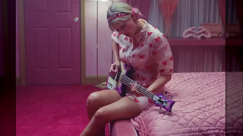 Swift sits on a pink mattress playing a pink guitar. She is wearing a light pink button-up dress covered in hot pink butterflies, a pink acrylic headband, and pink ribbons to pull back her hair into two low buns.