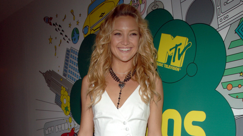 Kate Hudson in a white cropped vest with a black chunky necklace and her hair in loose waves with a bang pouf.