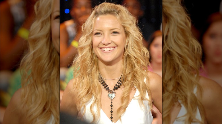 Kate Hudson in a white cropped vest with a black chunky necklace and her hair in loose waves with a bang pouf.