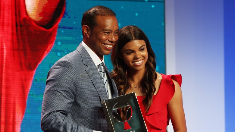 Sam Woods poses with her father with her hair down in loose waves with a middle part and a bright red sleeveless dress while attending the World Golf Hall of Fame Induction Ceremony in Florida in 2022.