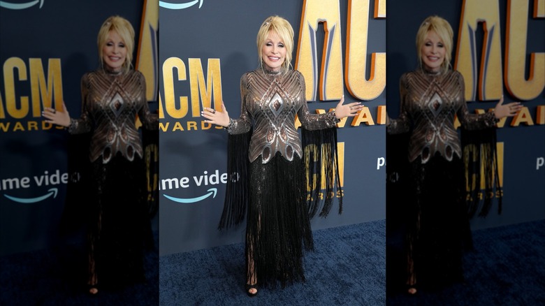 Dolly Parton with her arms out wearing a metallic dress with black fringe at the 2022 ACM Awards.