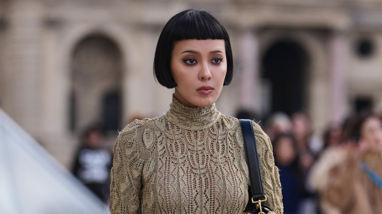 Kiwi Lee Han wears black hair in a bob haircut with bangs, a beige knit cable turtleneck sweater, a black and red plaid checkered mini skirt, a large yellow and green belt with argyle patterns a black leather bag, black leather embellished rhinestones ankle shoes, outside Louis Vuitton, during Paris Fashion Week - Womenswear Fall/Winter 2026/2027, on March 10, 2026