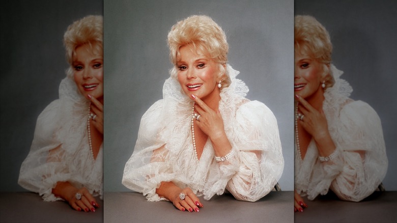 Eva Gabor poses with her hand by her face wearing a poofy lace blouse and long string of pearls.