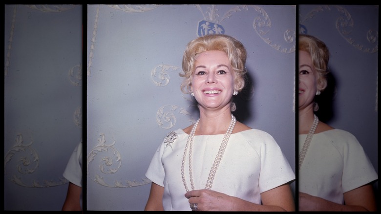 Eva Gabor smiles wearing a pearl necklace and a white dress against a blue wall.