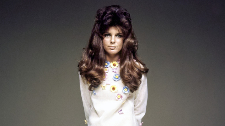Katharine Ross poses wearing a white dress with floral adornments and a voluminous hair piece.