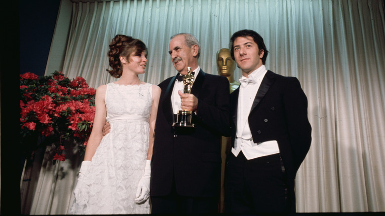 Katharine Ross standing alongside Burnett Guffey and Dustin Hoffman wearing a white lace dress and gloves.