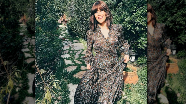 Erin Krakow is seen holding up the skirt of a flowy patterned dress in front of a scenic greenery background
