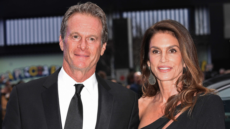 Rande Gerber wearing a black suit next to and Cindy Crawford in a black dress posing outside.