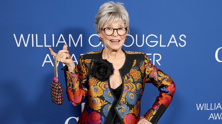 Rita Moreno posing at an event dangling a small purse on her finger and wearing a colorful outfit.
