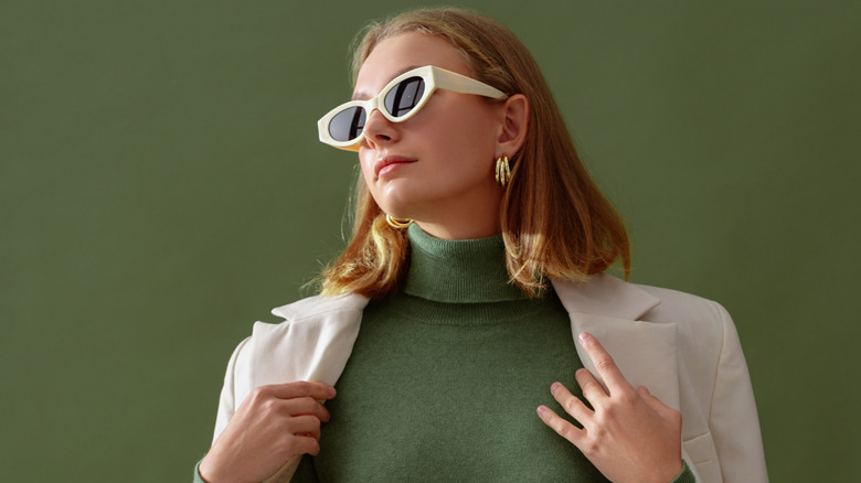 Woman wearing a green turtleneck top with a cream-colored jacket and white sunglasses in front of a green background.