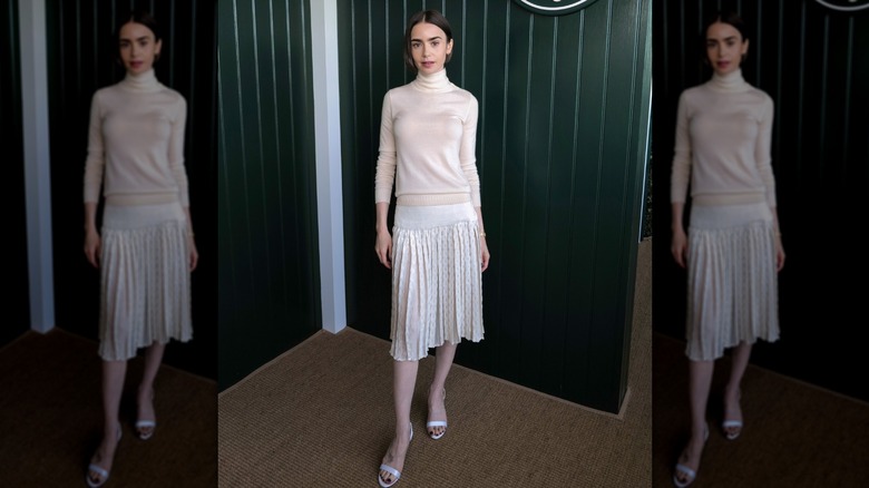 Lily Collins wearing a cream-colored turtleneck top and a white pleated skirt with white sandals at an event.