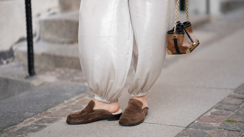 A cropped photo of a woman wearing backless suede loafers and baggy white pants.