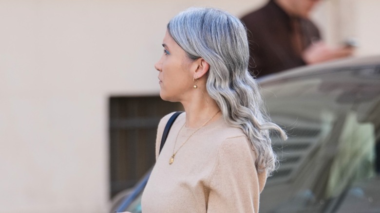 A woman with gray hair wearing a beige sweater and a pendant necklace looking to the side.