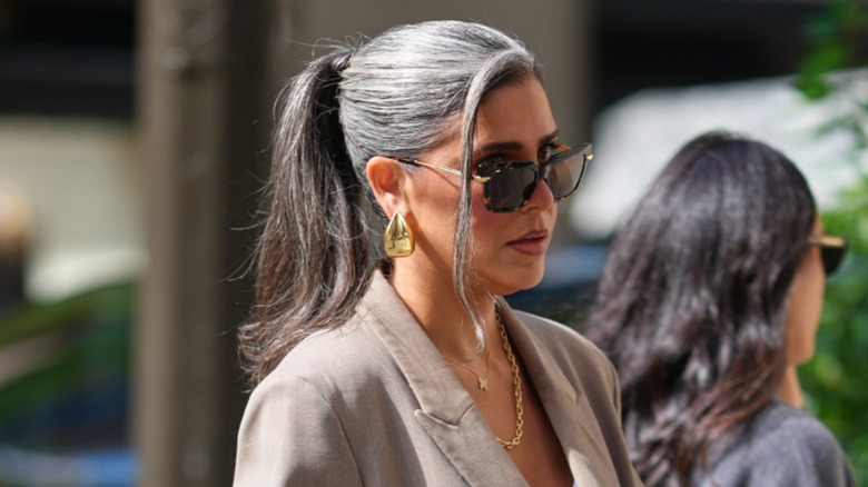 A woman with gray hair in a high ponytail wearing sunglasses, gold jewelry, and a gray blazer.