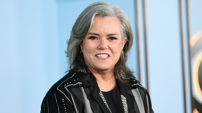 Rosie O'Donnell wears a black and grey striped shirt while posing in front of a blurred baby blue backdrop.