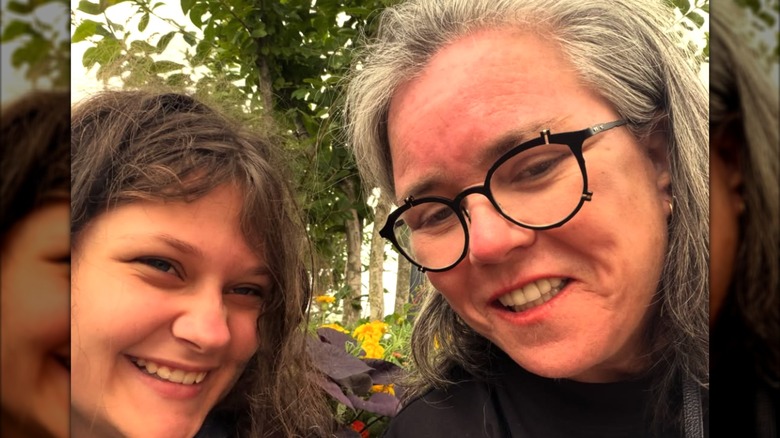 Rosie O'Donnell and her child Clay pose for a smiling selfie with greenery in the background.