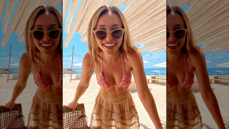 Camelia Somers wears sunglasses, a pink floral bikini top, and sheer tan skirt on the beach.