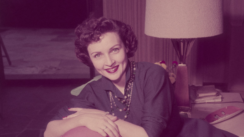 Betty White, US actress and comedian, smiling while sitting with her arms folded across a pillow on the backrest of a sofa in a living room, circa 1955
