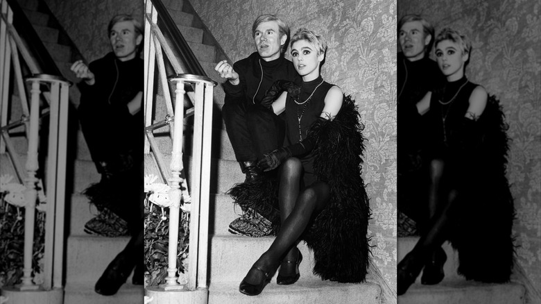Artist Andy Warhol wearing all black and Edie Sedgwick wearing a black outfit with a black feather boa, sitting on a staircase.