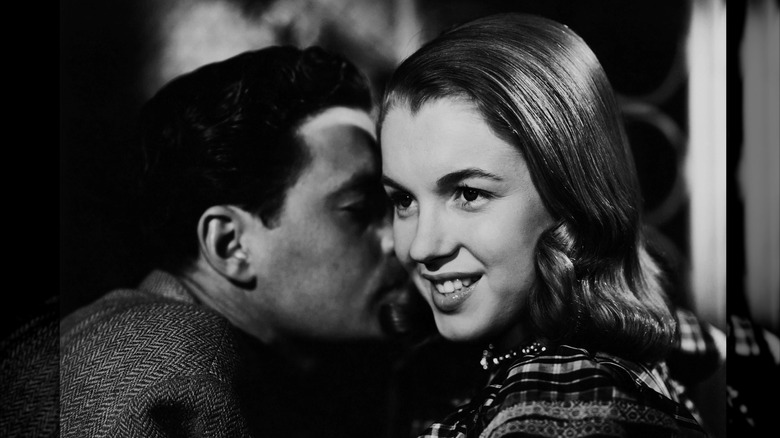 Black-and-white photo of Marilyn Monroe with sleek, brunette hair smiling while a man whispers in her ear.