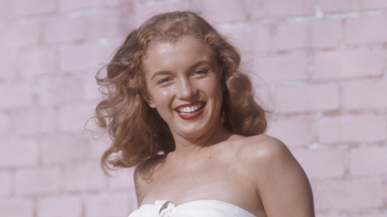 Marilyn Monroe posing in a white top with red lipstick and wavy strawberry-blond hair in front of a brick wall.