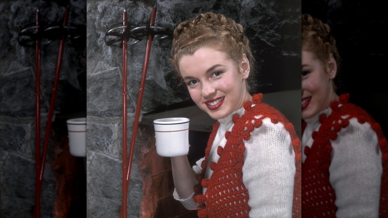 Marilyn Monroe posing in 1946 holding a mug with her hair in a braided updo wearing a red knit vest over a white sweater.