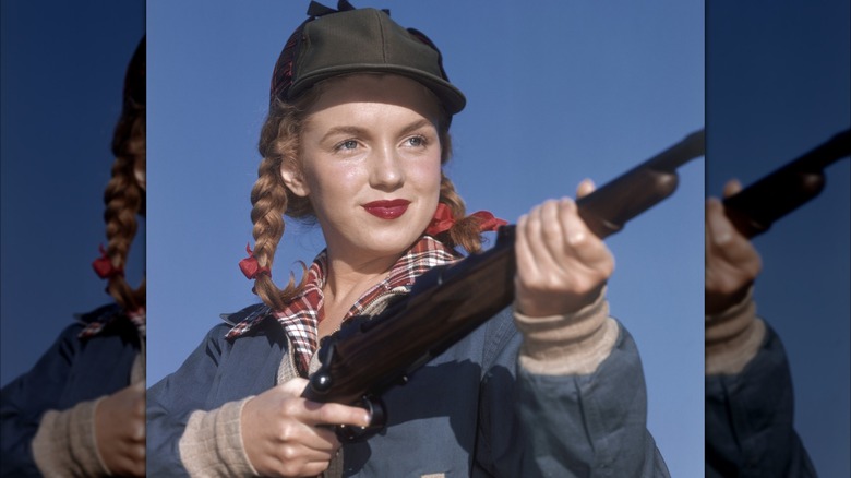 Marilyn Monroe posing in 1946 holding a rifle wearing a plaid shirt, hunting hat, and red lipstick with her hair in braids.