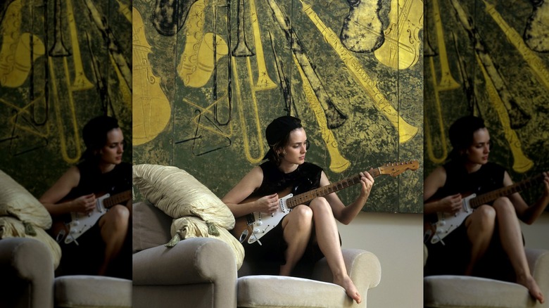 Winona Ryder playing guitar on a chair in front of a colorful art piece.