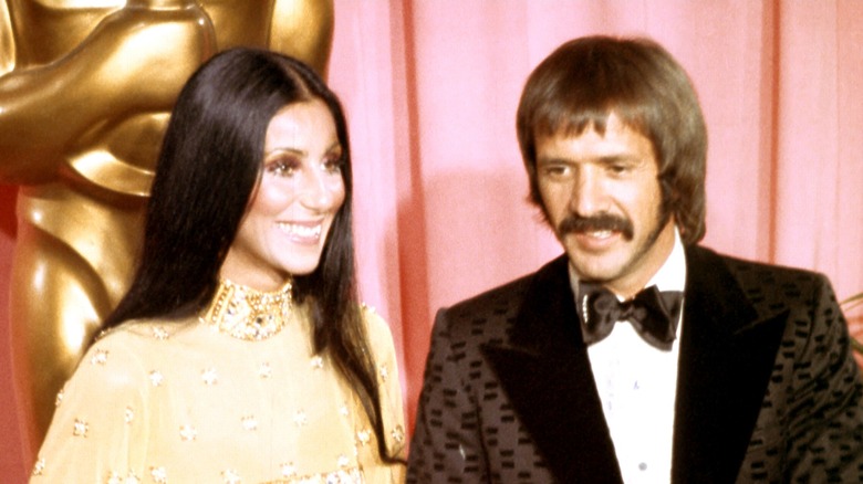 Cher wearing a gold gown with straight hair and Sonny Bono in a brown patterned suit pictured at the 1973 Academy Awards.