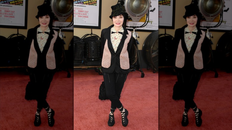 Toni Basil posing on the red carpet with her ankles crossed, wearing a black outfit with pink and white accents.