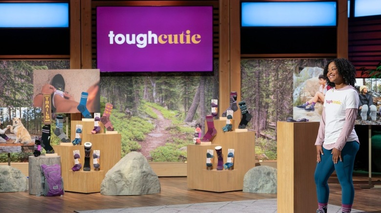 ToughCutie founder Brittany Coleman wears a white t-shirt with her company logo over a long-sleeve pink hooded shirt with jeans and her ToughCutie socks while pitching her business on Shark Tank, airing in January 2025.
