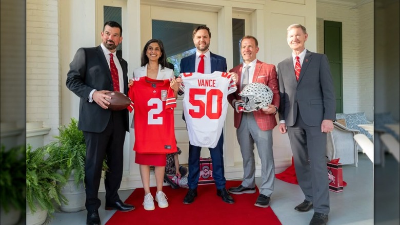 Usha Vance holds up a red football jersey next to her husband JD Vance and other men. She is wearing a white blazer, a long red skirt, and chunky white sneakers.