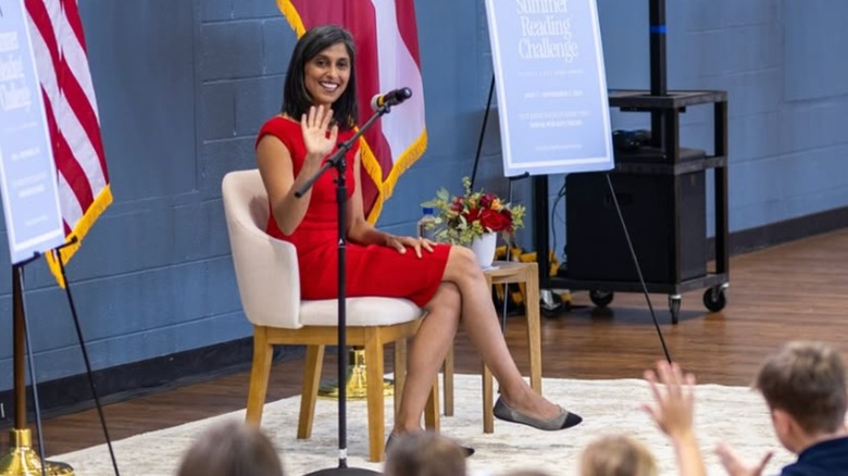 Usha Vance sits in front of a crowd of children. She is wearing a long red dress and ballet flats.