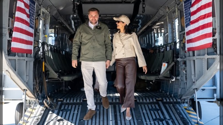 Usha Vance walks hand in hand with JD Vance on an aircraft carrier. She is wearing a cropped tan trenchcoat, brown leather pants, and white pointed toe heels.